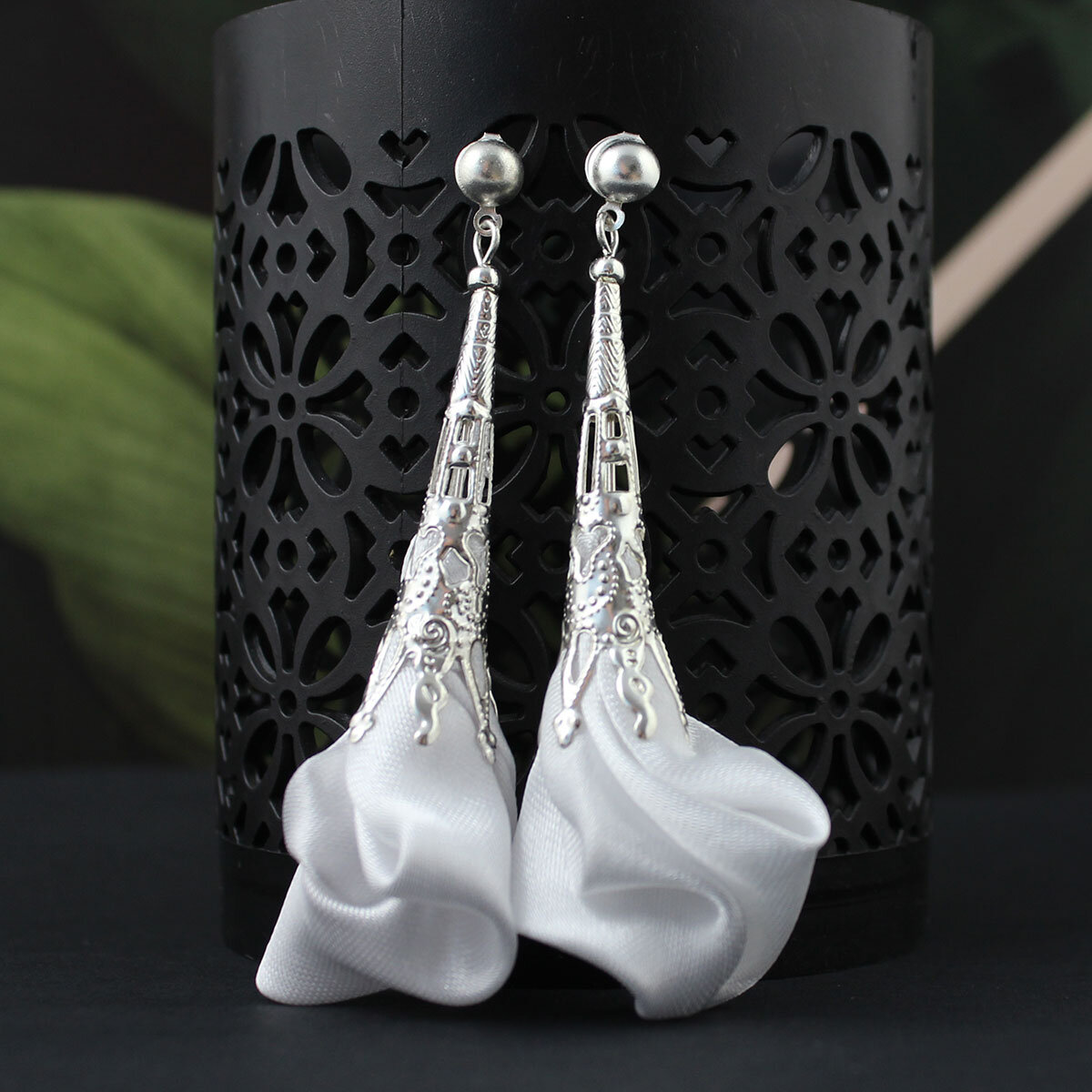 Długie, wiszące kolczyki w kształcie białych kwiatów uformowanych z tkaniny, zwieńczone ażurowym, filigranowym stożkiem w kolorze srebrnym na zapięciu typu sztyft