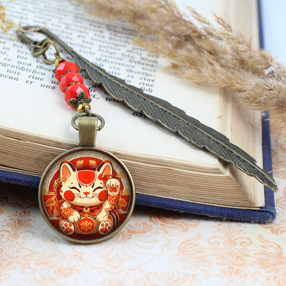 Zakładka do książki z antycznego brązu w kształcie pióra, z czerwonymi kryształkami i grafiką radosnego kota Maneki Neko pod szklanym kaboszonem.