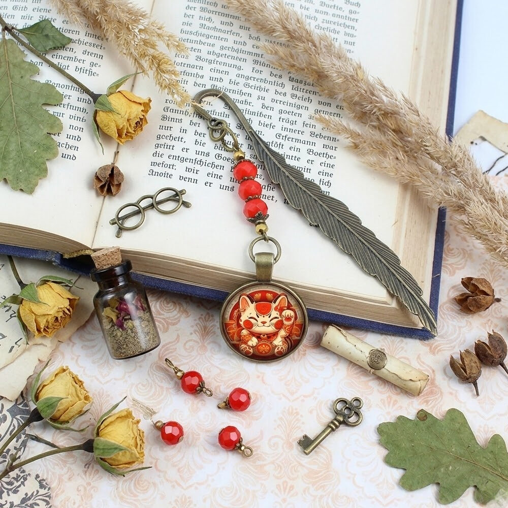 Zakładka do książki z antycznego brązu w kształcie pióra, z czerwonymi kryształkami i grafiką radosnego kota Maneki Neko pod szklanym kaboszonem.