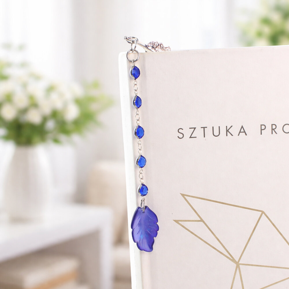 Cienka i lekka metalowa zakładka do książki w kształcie pióra, zdobiona wiszącą zawieszką z szafirowych szklanych kryształków i niebieskiego akrylowego listka, w ozdobnym pudełku.