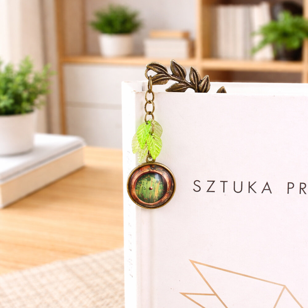 Zakładka do książki z antycznego brązu z zawieszką w formie zielonych listków i grafiką drzwi do domu Hobbita pod szklanym kaboszonem.