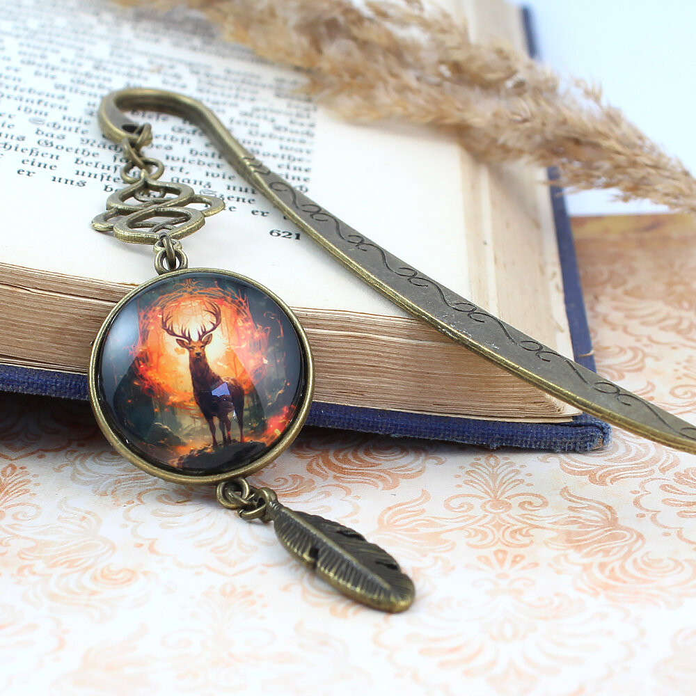 Zakładka do książki z antycznego brązu z ozdobnym falistym trzonem, zawieszką z piórkiem i ilustracją jelenia w ognistym lesie pod kaboszonem.