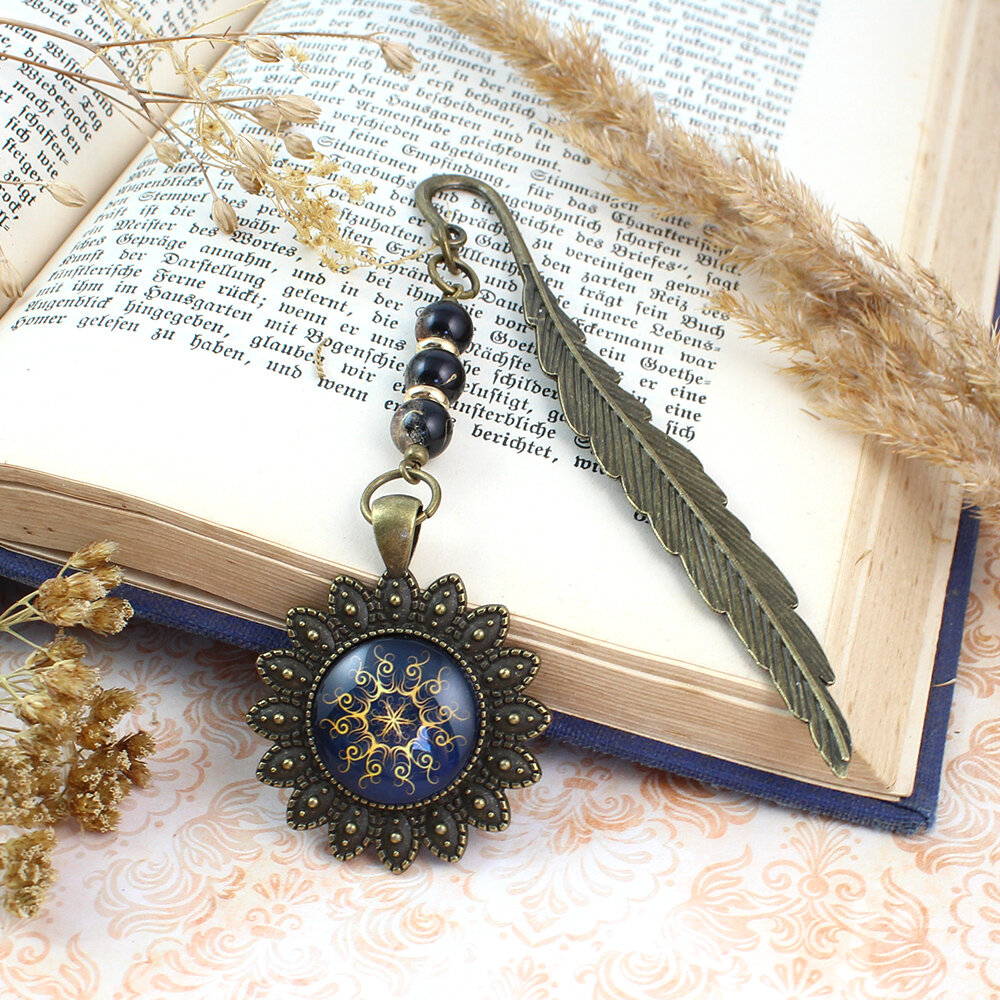 Zakładka do książki z antycznego brązu w kształcie pióra, z ozdobnymi koralikami oraz słoneczną, złotą mandalą na granatowym tle w oprawie z płatków