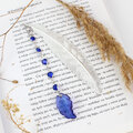 Cienka i lekka metalowa zakładka do książki w kształcie pióra, zdobiona wiszącą zawieszką z szafirowych szklanych kryształków i niebieskiego akrylowego listka, w ozdobnym pudełku.