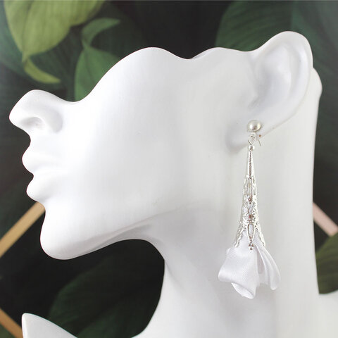 Długie, wiszące kolczyki w kształcie białych kwiatów uformowanych z tkaniny, zwieńczone ażurowym, filigranowym stożkiem w kolorze srebrnym na zapięciu typu sztyft