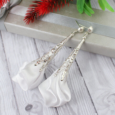 Długie, wiszące kolczyki w kształcie białych kwiatów uformowanych z tkaniny, zwieńczone ażurowym, filigranowym stożkiem w kolorze srebrnym na zapięciu typu sztyft