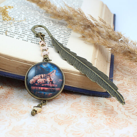 Zakładka do książki z antycznego brązu z zawieszką w kształcie gwiazdek oraz ilustracją śpiącego kota na stosie książek pod szkiełkiem.