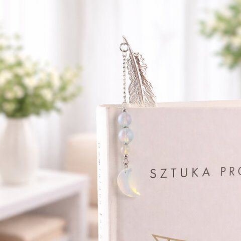 Subtelna metalowa zakładka do książki w formie piórka, zdobiona wiszącą zawieszką z opalitu w kształcie księżyca, umieszczona w pudełku prezentowym.